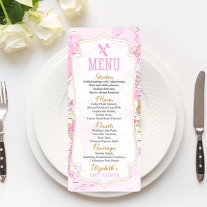 Puede incluir: Un menú de baby shower rosa y dorado con un fondo de mapa y un pequeño avión gráfico. El menú incluye entrantes, platos principales, postres, bebidas y el texto "Baby Shower de Elizabeth".