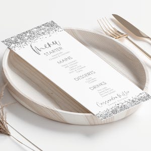 Può includere: Un biglietto di menu bianco con dettagli glitterati argentati, con antipasti, piatti principali, dessert e bevande. Poggia su un piatto di legno, accompagnato da posate dorate. L'erba ornamentale secca è visibile sullo sfondo.