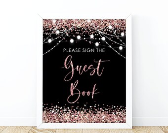 May include: A framed sign with a black background and rose gold glitter accents. The text "PLEASE SIGN THE Guest Book" is displayed in white and rose gold script. White string lights are at the top of the sign.