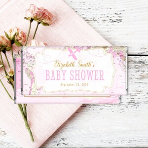 May include: Pink and gold baby shower chocolate bar wrapper with a world map design and the text "Elizabeth Smith's Baby Shower September 24, 2023".