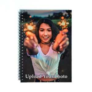 May include: A spiral bound notebook with a black cover featuring a photo of a person smiling and holding sparklers. The text "Upload Your Photo" is printed on the cover.