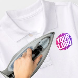 May include: A white polo shirt with a collar and a button placket. A circular graphic with a pink and purple gradient border contains the white text YOUR LOGO. A grey and black steam iron is positioned over the fabric of the white polo shirt.