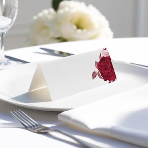May include: A white place card with a red rose and green leaves printed on it. The place card is folded in half and is sitting on a white plate.