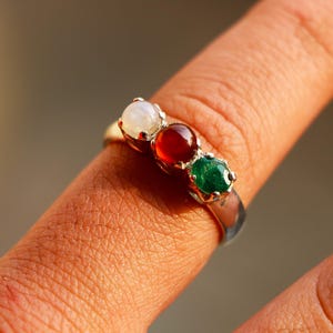May include: A silver ring featuring three gemstones: a white moonstone, a red garnet, and a green emerald. The ring is displayed on a finger, highlighting the colours and design of the jewellery.