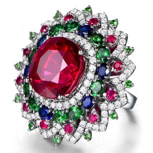 Puede incluir: Un anillo de tono plateado con una gran piedra preciosa roja central de corte ovalado, rodeada por un halo floral de varias capas. El diseño incluye piedras preciosas redondas más pequeñas en rojo, verde y azul, intercaladas con numerosos cristales blancos en pavé.