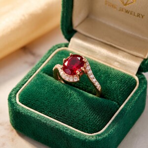 May include: A rose gold ring featuring a large oval-cut deep red gemstone at the centre, surrounded by a halo of small clear stones. The band is also set with a row of small clear stones, presented in a dark green velvet jewellery box.