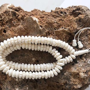 May include: A string of off-white prayer beads featuring small, disc-shaped beads with carved vertical grooves. The beads are strung on a thin cord that terminates in a larger, smooth, elongated bead and two smaller, matching end pieces.