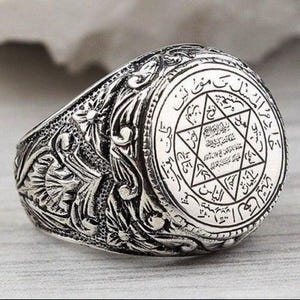 May include: A silver-toned ring featuring an intricate, raised floral pattern along the band. The circular face displays a central six-pointed star surrounded by detailed Arabic script, with additional text inscribed within the star's geometric lines.
