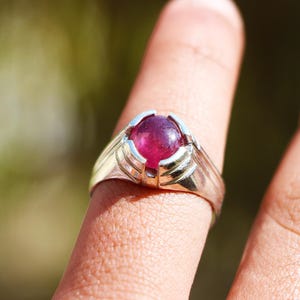 May include: A silver ring with a large, round, deep pink gemstone. The ring's setting has a detailed design that cradles the stone. The ring is shown on a finger against a blurred green background.