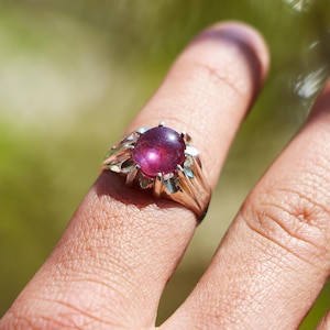 May include: A silver ring featuring a large, oval, purplish-red gemstone. The ring has an intricate, ornate setting with a floral design. The ring is on a finger, with a blurred green backdrop.