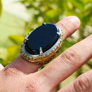 May include: An oval black gemstone ring with a silver and gold filigree setting. The ring is worn on a finger, showcasing the polished black stone and intricate metalwork. The ring's design features a gold band with silver accents.