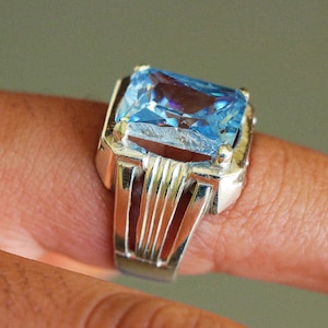 May include: A silver ring with a large, rectangular, light blue gemstone. The ring has a detailed, vertical bar design on the sides of the band. The gemstone is faceted, catching the light and creating a sparkling effect. The ring is on a finger.