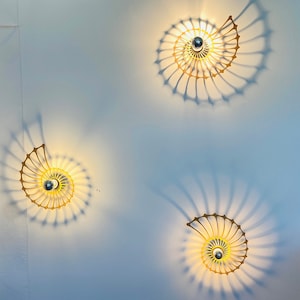 Puede incluir: Tres apliques de pared con diseño de concha de nautilus. Los apliques son de color dorado y proyectan una sombra en espiral sobre la pared blanca. Cada aplique tiene una fuente de luz central.