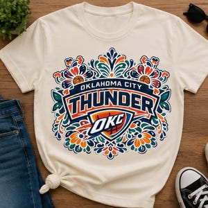 Op de afbeelding: Crèmekleurig T-shirt met het Oklahoma City Thunder-logo, versierd met bloemen- en bladerenmotieven. Het ontwerp bevat de teamnaam en "OKC" in een levendig kleurenpalet. Het shirt wordt gecombineerd met een blauwe jeans en zwarte sneakers.