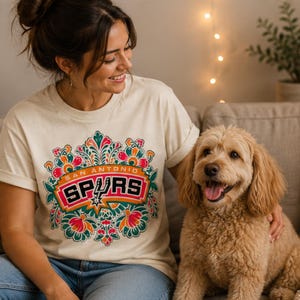 May include: Cream-colored t-shirt featuring a colorful floral design with the text "SAN ANTONIO SPURS" in a red and orange banner. The shirt is worn by a person sitting on a couch next to a light brown dog.
