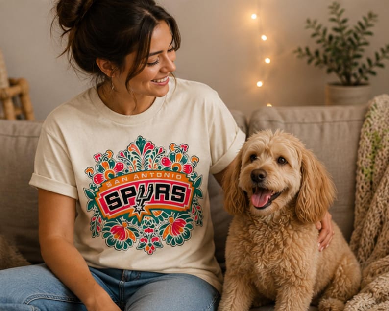 May include: Cream-colored t-shirt featuring a colorful floral design with the text "SAN ANTONIO SPURS" in a red and orange banner. The shirt is worn by a person sitting on a couch next to a light brown dog.
