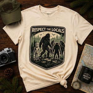 May include: Cream-colored t-shirt with a graphic featuring an alien, Bigfoot, and a winged creature, with the text "Respect the Locals." The design is set against a forest backdrop. Additional items include a green cap, a vintage camera, and a compass.