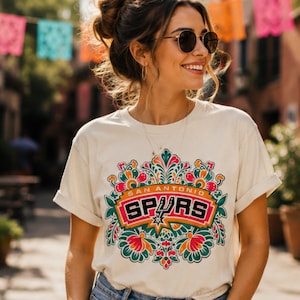 May include: Cream-colored t-shirt featuring a colorful floral design with the text "SAN ANTONIO SPURS" in a rectangular emblem. The design includes a star and is surrounded by orange, green, and red floral elements.