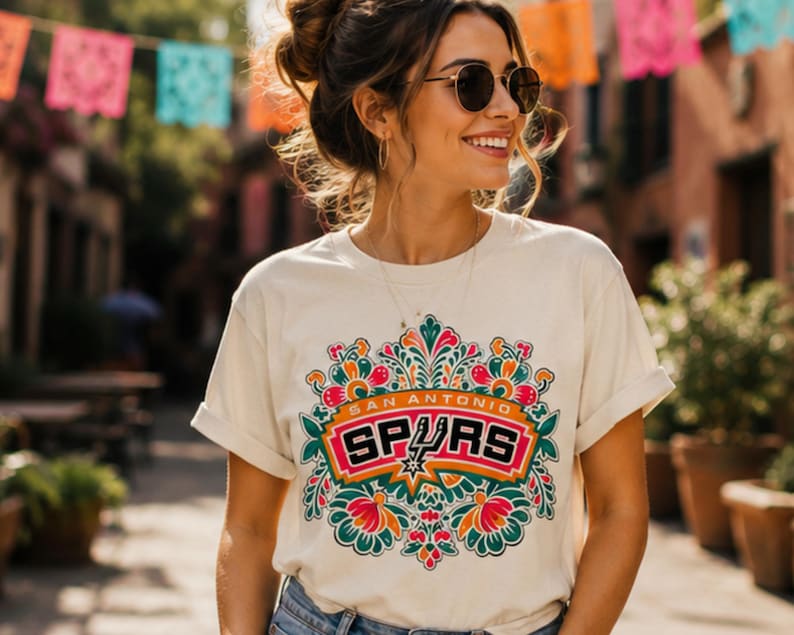 May include: Cream-colored t-shirt featuring a colorful floral design with the text "SAN ANTONIO SPURS" in a rectangular emblem. The design includes a star and is surrounded by orange, green, and red floral elements.
