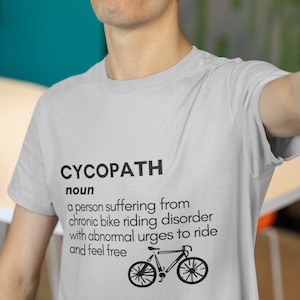 Può includere: Una maglietta grigia con la parola "CYCOPATH" definita come "una persona che soffre di un disturbo cronico della guida in bicicletta con voglie anomale di andare in bicicletta e sentirsi libera". Sotto il testo c'è un'illustrazione in bianco e nero di una bicicletta.