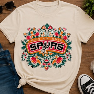 May include: Cream-colored t-shirt featuring the San Antonio Spurs logo with floral and geometric designs in red, green, and orange. The shirt is paired with blue jeans and black and white sneakers.