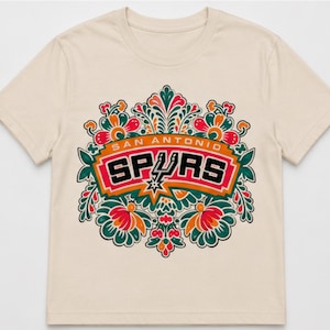 May include: Cream-colored t-shirt featuring the San Antonio Spurs logo. The logo is set within a floral design with red, orange, green, and white accents. The text "SAN ANTONIO SPURS" is displayed above the logo.