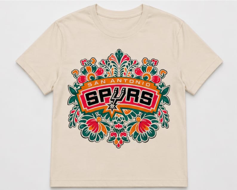 May include: Cream-colored t-shirt featuring the San Antonio Spurs logo. The logo is set within a floral design with red, orange, green, and white accents. The text "SAN ANTONIO SPURS" is displayed above the logo.