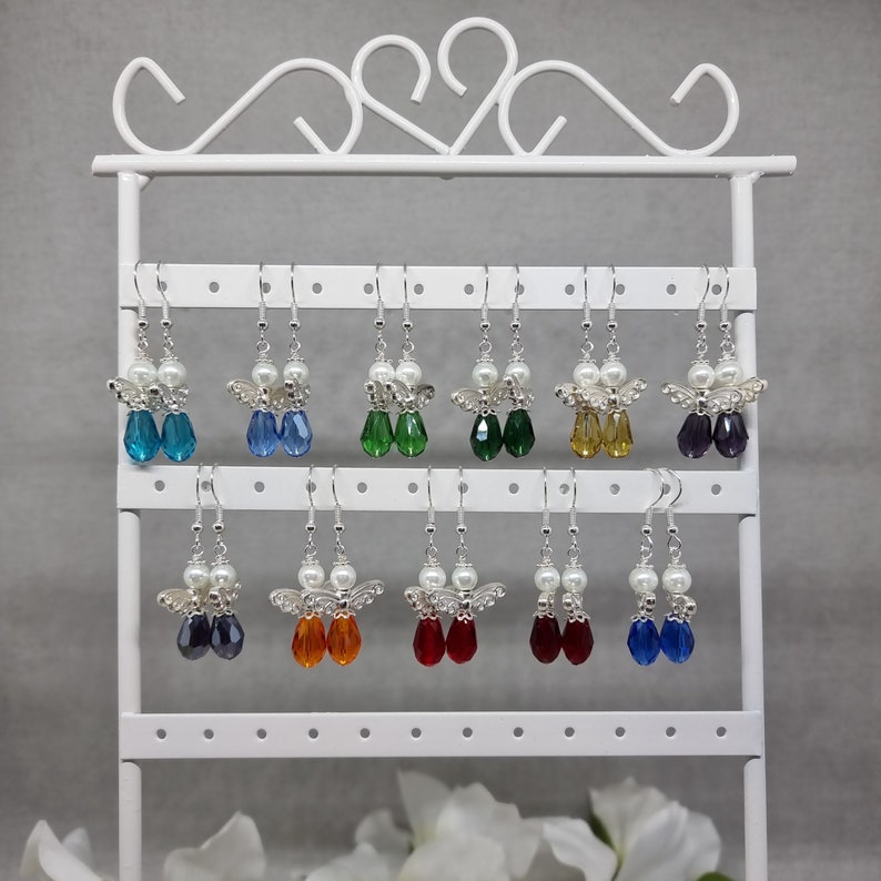 May include: A display of silver angel wing earrings with crystal beads in various colours, including blue, green, red, orange, and purple. The earrings are displayed on a white metal stand.