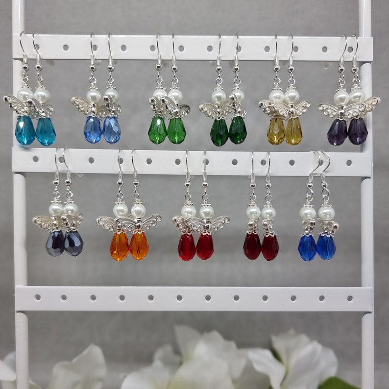 May include: A display of 12 pairs of angel wing earrings with crystal drops in various colours, including blue, green, red, orange, and purple. The earrings are displayed on a white earring stand.