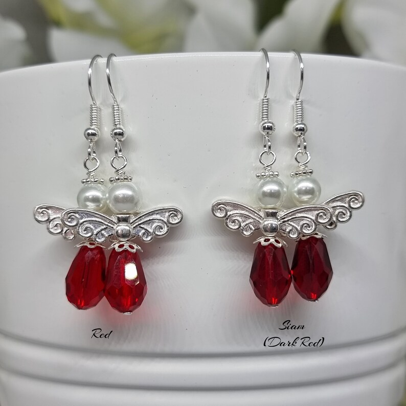 May include: Two pairs of silver angel dangle earrings with red crystal beads. The earrings on the left have red beads and the earrings on the right have darker red beads. The text "Red" is below the left earrings and the text "Siam (Dark Red)" is below the right earrings.