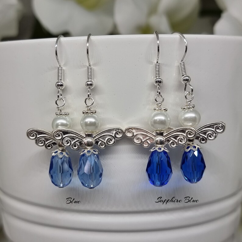 May include: A pair of silver angel wing earrings with a pearl bead and a blue crystal drop. The earrings are shown in two colours: blue and sapphire blue.