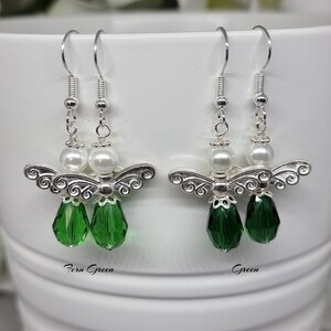 May include: A pair of silver angel earrings with green crystal drops. The earrings have a white pearl bead above the angel. The earrings are labelled "Fern Green" and "Green".