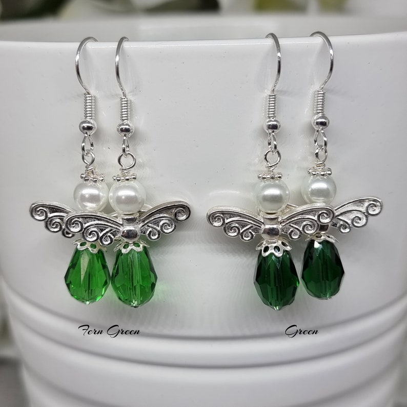 May include: A pair of silver angel earrings with green crystal drops. The earrings have a white pearl bead above the angel. The earrings are labelled "Fern Green" and "Green".