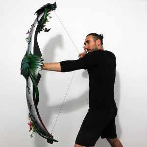 May include: A person is holding a green and silver bow with floral accents. The bow is made of wood and has a string attached to it. The person is wearing a black shirt and black shorts.
