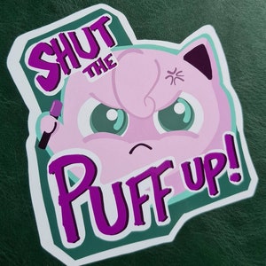 May include: A sticker featuring a pink cartoon character with an angry expression and the text "SHUT THE PUFF UP!" in purple. The character has green eyes and a small black microphone.