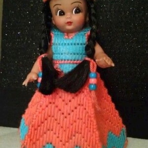 May include: A doll dressed in a Native American-inspired outfit. The doll has black braids, a blue and orange headdress with feathers, and a dress with a zig-zag pattern. The doll's dress is orange with blue accents.