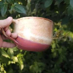 Könnte beinhalten: Ein Keramikbecher mit zweifarbigem Design, bestehend aus einem gesprenkelten cremefarbenen oberen Teil und einer soliden dunkelrotbraunen Basis. Der Becher hat einen breiten, abgerundeten Körper und einen seitlich angebrachten, geschwungenen Henkel.