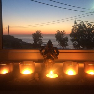 May include: A serene scene featuring a window view of a sunset over the ocean. In the foreground, lit candles in glass holders and a small statue of a deity create a peaceful ambiance. The warm glow of the candles contrasts with the cool tones of the sky.