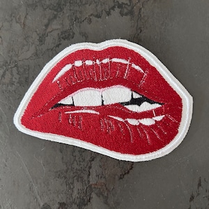 Op de afbeelding: Rode geborduurde lippen met witte tanden en een zwarte omtrek. De lippen zijn lichtjes geopend, waardoor de tanden zichtbaar zijn. De borduurwerk is op een witte stoffen achtergrond.