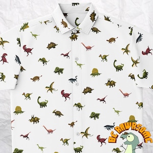 Op de afbeelding: Wit overhemd met korte mouwen en knopen met een herhalend patroon van kleurrijke gepixelde dinosaurussen. Het overhemd heeft een knoopsluiting en een zakje op de linkerborst. De tekst "BE RAWESOME" is afgedrukt in de rechterbenedenhoek van het overhemd.