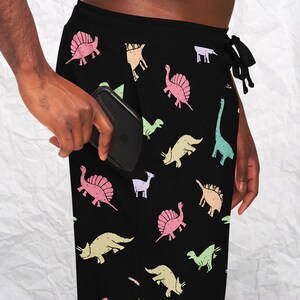 May include: Black lounge pants with a colorful dinosaur pattern. The pants have a drawstring waist and a pocket. A smartphone is in the pocket. The dinosaurs are in shades of pink, green, yellow, and blue.
