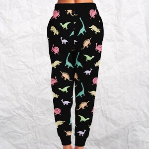 May include: Black sweatpants with a colorful dinosaur pattern. The dinosaurs are in various pastel colors, including pink, yellow, green, and blue. The pants have a drawstring waist and pockets.