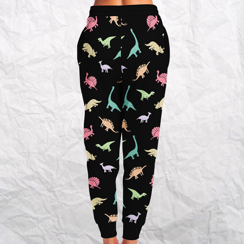 May include: Black sweatpants with a colorful dinosaur pattern. The dinosaurs are in various pastel colors, including pink, yellow, green, and blue. The pants have a drawstring waist and pockets.