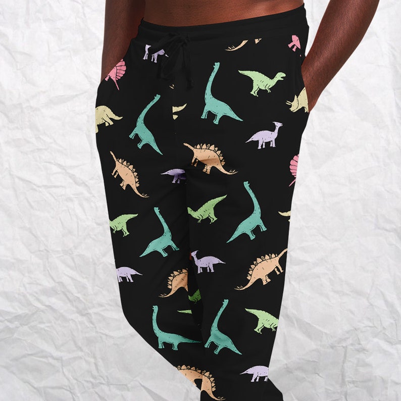May include: Black sweatpants with a colorful dinosaur pattern. The dinosaurs are in shades of green, blue, orange, pink, and purple. The pants have a drawstring waist and pockets.