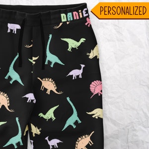 May include: Black sweatpants with a colorful dinosaur pattern. The dinosaurs are in pastel colors, including blue, green, pink, and yellow. The waistband has the name "DANIE" in rainbow letters. The word "PERSONALIZED" is in a yellow box.