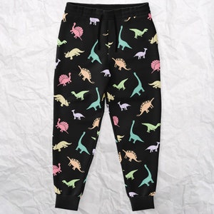 May include: Black sweatpants with a colorful dinosaur pattern. The dinosaurs are in shades of pink, yellow, green, and blue. The pants have a drawstring waist and elastic cuffs.
