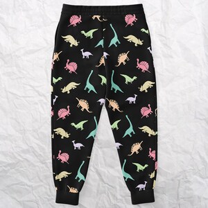May include: Black sweatpants with a colorful dinosaur pattern. The dinosaurs are in various pastel colors, including pink, yellow, green, and blue. The sweatpants have a drawstring waist and ribbed cuffs at the ankles.