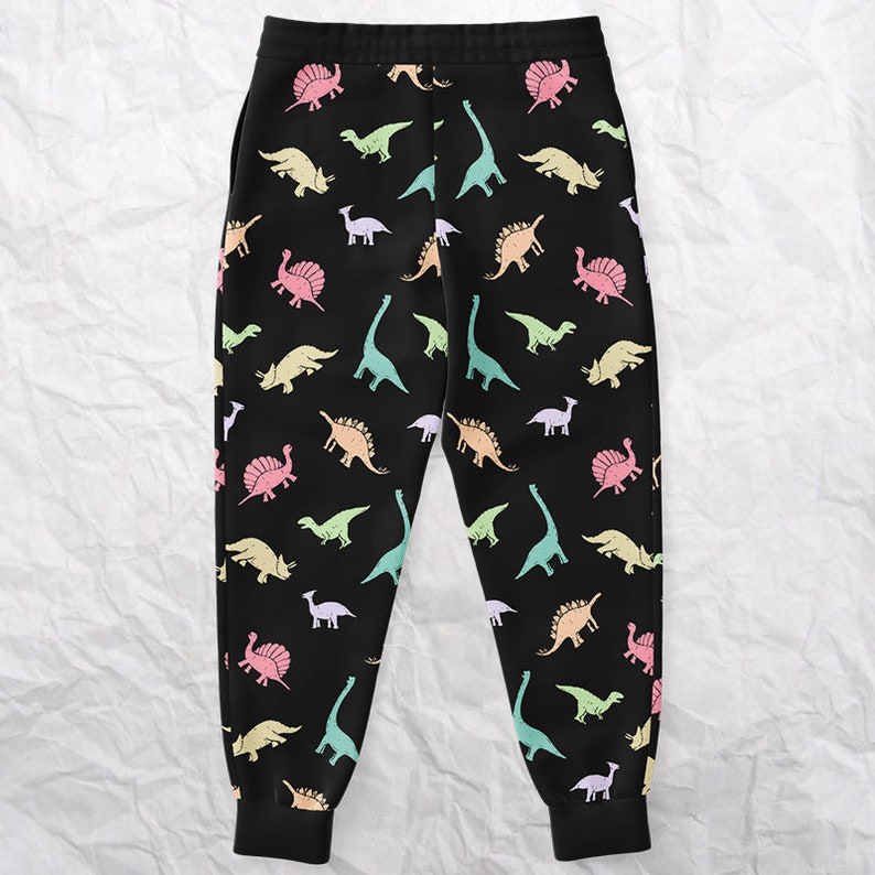 May include: Black sweatpants with a colorful dinosaur pattern. The dinosaurs are in various pastel colors, including pink, yellow, green, and blue. The sweatpants have a drawstring waist and ribbed cuffs at the ankles.