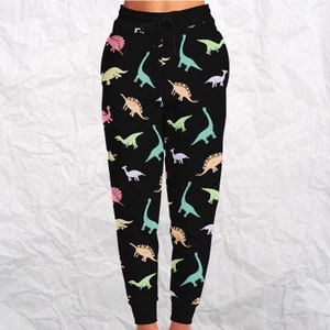 May include: Black sweatpants with a repeating pattern of pastel-colored dinosaurs. The dinosaurs are in various poses and species, including Brontosaurus, Stegosaurus, and Triceratops. The pants have a drawstring waistband.