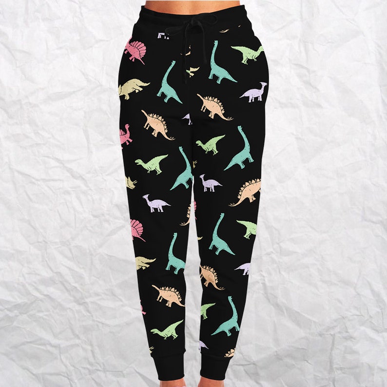 May include: Black sweatpants with a repeating pattern of pastel-colored dinosaurs. The dinosaurs are in various poses and species, including Brontosaurus, Stegosaurus, and Triceratops. The pants have a drawstring waistband.